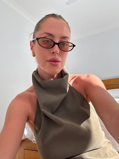 A woman with blonde hair pulled back and dark-rimmed glasses poses. She is wearing a sleeveless, olive green top with a draped neckline. The photo is taken from a low angle, showing her face, shoulders, and part of her chest. Light shines on her arms and the top. A partial view of a wooden frame or headboard is visible in the background, along with a white wall.