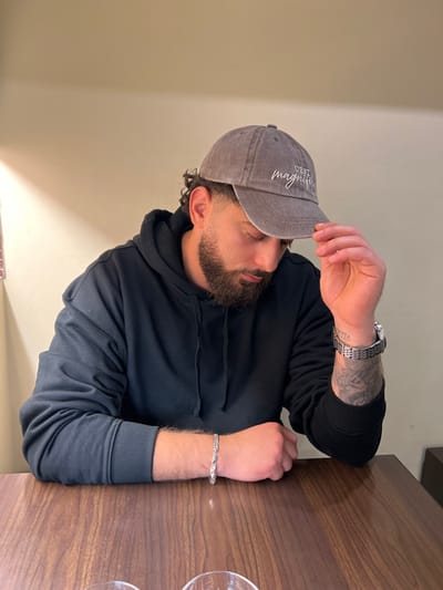 A male subject is depicted seated at a dark wooden table in an indoor establishment located in Chevilly-Larue, France. The individual wears a dark grey or black hooded sweatshirt and a faded grey baseball cap with "C'EST magnifique" embroidered in white on the front. His head is bowed, looking downwards, and his right hand is resting on the brim of the cap. He possesses a full dark beard and curly dark hair visible around his ears. A silver-toned chain bracelet is present on his left wrist. On his right wrist, a silver-toned metallic watch with a link band and a visible face featuring Roman numerals is worn, above which a dark tattoo is present on the inner forearm. Two clear glass tumblers are partially visible in the lower foreground on the table. The background consists of a plain, light-colored wall.