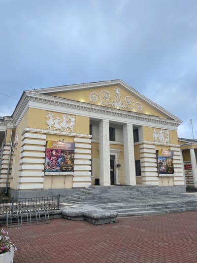 The image shows the facade of the "Energiya" cinema in Zavolzhye, Russia. The building has a classical architectural style with a yellow exterior, white columns, and decorative relief sculptures on its facade. Two movie posters are displayed on the left and right sides of the entrance. The entrance is approached by a wide set of concrete stairs. In front of the building, there is a red brick pavement and a metal bicycle rack. A white planter with pink and white flowers is visible in the lower left corner. The sky above is overcast.