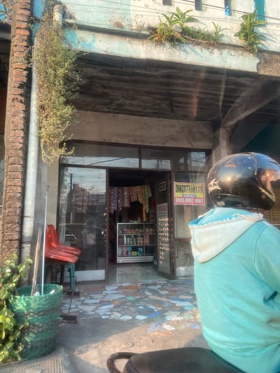 The image captures a street-level view of a shop front in Jekulo, Indonesia. Sunlight casts strong shadows across the scene. In the foreground, a person on a motorbike wears a black helmet and a turquoise hooded jacket. The shop itself has a glass entrance and displays various goods on shelves. A sign on the shop reads "DIKONTRAKKAN" with a phone number. Red plastic chairs are visible outside the shop entrance, and a large green woven basket sits to the left. The building's facade features peeling paint and vegetation growing from upper ledges. The ground in front of the shop is tiled with a mosaic pattern.