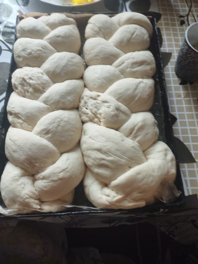 The image shows a baking pan filled with braided dough. The dough is a light, pale color and appears to be raw, not yet baked. There are multiple braids in the pan, arranged in two rows. The pan itself is dark and rectangular, with visible edges. The background shows a checkered tablecloth and a dark object with a textured surface, possibly a container. There is no indication of people or specific locations in Ciuciulea, Moldova, in the image.