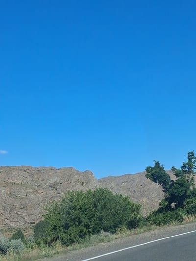The image depicts a scenic view of a rugged, rocky mountainside with scattered green vegetation. A paved road with a white line marking runs diagonally across the lower right portion of the frame. The sky is a clear, vibrant blue with a small wisp of cloud visible on the far left. The landscape suggests a rural or natural setting, likely outside of a dense urban area. The specific location in Malishka, Armenia, would imply this is a view of the natural geography surrounding the village.
