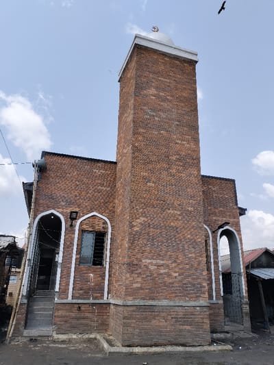 The image displays a mosque constructed primarily of reddish-brown brick, situated in Oworonshoki, Nigeria, under a partly cloudy sky. The building features a prominent, rectangular minaret on the right, topped with a light-colored dome and a golden crescent moon and star finial. A dark bird is visible flying in the upper right quadrant of the sky.
The main structure of the mosque has a dark roofline and multiple arched openings framed with white trim. On the left side of the building, there are two distinct arched openings. The leftmost opening functions as an entrance with several steps leading up to it, featuring metal grating at the base and a dark interior visible. Above this entrance, a grey horn speaker is mounted on the wall, partially obscured by power lines and wires. Adjacent to this entrance is another arched opening containing a window with horizontal security bars and a dark, possibly reflective, pane. A black rectangular light fixture is mounted on the brick wall between these two arches.
The ground directly in front of the mosque appears to be dirt or unpaved, with a raised concrete curb segment separating it from the building's base. To the far left and right of the mosque, rudimentary structures with corrugated metal roofs and weathered facades are partially visible, suggesting a dense urban or semi-urban setting. No individuals or direct interactions are visible in the scene.