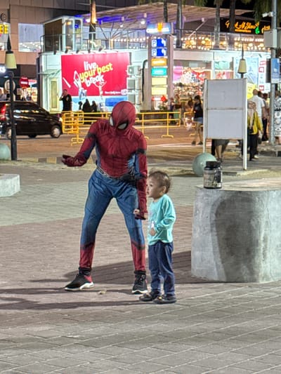 The scene depicts a nighttime urban environment, identified as Cenang Beach, Malaysia, characterized by artificial lighting. In the foreground, a person costumed as Spider-Man stands to the left of a young child. The Spider-Man costume is red and blue, covering the entire body, including a mask, with black athletic shoes. The figure's right hand is open, palm up, extended towards the child, while the left arm is bent.

To the right of Spider-Man, a toddler-aged child stands, wearing a light blue long-sleeved shirt, dark blue pants, and dark sandals. The child faces right, away from Spider-Man, with an open mouth and hands loosely clasped in front.

The ground is paved with light-colored interlocking bricks. On the far right, a grey concrete block supports a dark bottle or container. In the background, a busy street and commercial area are visible. A black vehicle is partially seen on the left. A prominent red LED billboard displays "live your best break" in white text. Multiple storefronts, glowing signs, and streetlights illuminate the background. Yellow crowd control barriers are positioned along the street. Several blurred figures of pedestrians are present in the distance.