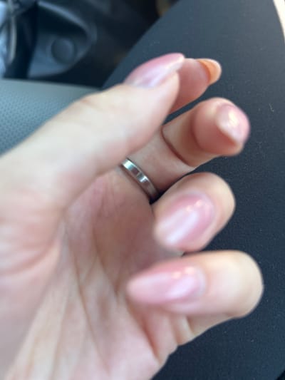 A close-up, slightly blurred, over-the-shoulder view of a person's hand with light pink fingernails is presented. The hand is positioned over a dark, textured surface, possibly clothing. A silver ring is visible on the ring finger, featuring a simple, unadorned band. The background is also out of focus, showing hints of a car interior with gray, textured upholstery and a black, circular object. The image does not provide any discernible landmarks or context to suggest a specific location within the city of Everett, United States of America.