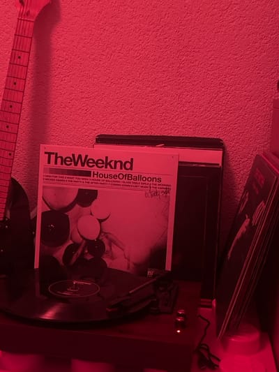 The image shows a record player with The Weeknd's "House of Balloons" vinyl album cover. The track list is visible, including songs like "High for This," "What You Need," and "Wicked Games." The vinyl is spinning on the turntable. A portion of a guitar neck is visible in the foreground, suggesting a musical setting. Stacks of records are visible behind the record player. The overall color scheme of the image is predominantly red.