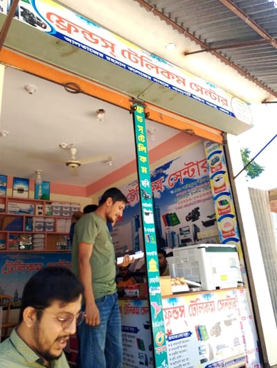 The image shows the interior of a small storefront in Pangasia, Bangladesh, located at coordinates 22.686 latitude, 90.618 longitude. The store is called "Kurus Telecommunication," and its facade is decorated with colorful signage and banners. Inside, a man in a green shirt and jeans stands by a counter. He appears to be talking to another man, who is partially obscured from view. Behind the counter is a large printer and a small counter with a variety of printed materials. The ceiling is white and has several light fixtures, and the walls are painted in light colors. There is a ceiling fan in the background. The storefront is bright and well-lit, with the sun shining through the front windows. The store appears to be a popular local establishment, with a variety of items for sale and a bustling atmosphere.