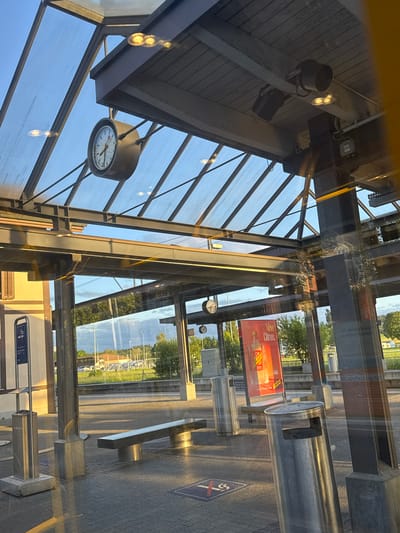 This image captures the interior of a modern building, likely a public transport stop or waiting area, looking out through large, reflective glass panels. The scene is brightly lit by natural light, suggesting a clear day.
The interior architecture features a ceiling constructed with horizontal wooden slats and robust metal beams. Several dark spotlights are mounted on these beams, and light reflections are visible on the glass. A prominent round clock with a dark frame, white face, and black hands hangs from the ceiling, displaying the time as approximately 10:10. More metal structural elements and glass sections complete the indoor framework.
Through the extensive glass, a vibrant outdoor environment is visible. Green trees and grass form a backdrop, indicating a natural setting beyond the immediate vicinity. A large, rectangular billboard, primarily red, is clearly visible, advertising "New Citrus. Schweppes." with smaller, less distinct text also present. The ground outside appears paved, with various metallic public amenities including cylindrical bins or ashtrays and a rectangular bench. A blue square sign on the paved ground displays a white circle with a crossed-out cigarette, signifying a "no smoking" zone. Another blue sign with a smoking symbol is affixed to a vertical surface, possibly indicating an ashtray.
The glass panels exhibit strong reflections, superimposing elements of the interior architecture onto the exterior view and creating a layered, somewhat abstract visual effect. The overall impression is of a calm, static scene under bright sunlight, with no people present. The time of day appears to be late morning, given the position of the sun and the clock reading. The context indicates the location is Fehraltorf, Switzerland.