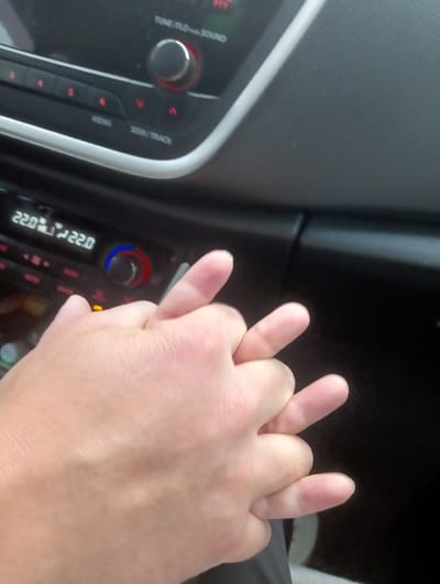 The image shows a close-up of two hands intertwined in a car's interior. The hands are clasped together, with fingers interlocked. In the background, the car's dashboard is visible, featuring a car stereo system with buttons and a digital display showing "22.0" degrees Celsius. The environment appears to be inside a vehicle, possibly in motion or parked. There is no information or visual cues within the image to determine the specific location of Nizhnetroitskiy, Russia. The focus is on the intimate gesture of the hands.