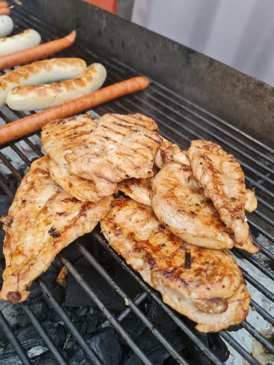 In Garbsen, Germany, several pieces of grilled chicken breast are piled on a black metal grate over hot coals. The chicken is golden brown with visible grill marks. In the background, several sausages are also grilling on the same grate. The scene depicts a barbecue or outdoor cooking event.