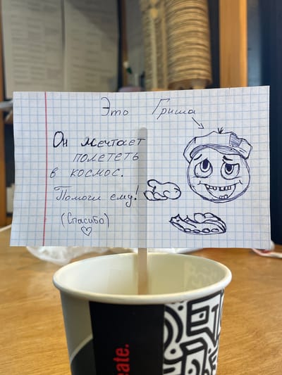 A note written on graph paper is attached to a wooden stick inserted into a paper coffee cup. The note reads: "Это Гриша. Он мечтает полететь в космос. Помоги ему! (Спасибо) ♡". A drawing of a cartoon face with a hat and an open mouth with visible teeth is on the right side of the note. The coffee cup has a black and white patterned design. The background shows blurred interior elements, possibly bookshelves or wall decor. The scene is indoors, likely in a cafe or similar establishment, and the note is a playful request for assistance, referencing someone named Grisha. There is no indication of the location being in the city of Shiryaevo, Russia.