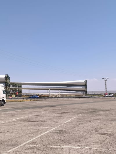 In Corella, Spain, a load of three large, white wind turbine blades is stacked horizontally on a multi-level rack system. The blades are the dominant objects in the scene, stretching across a significant portion of the frame. A white semi-truck, presumably used for transport, is partially visible on the left side of the image.

The scene appears to be an outdoor staging area or a transport hub, indicated by the paved surface with visible road markings in the foreground and a highway barrier separating the staging area from a road. Several vehicles are present on the road, including a blue car driving alongside the turbine blades and a white SUV parked on the right. A tall, lattice-like transmission tower stands in the background, with power lines extending across the sky.

The sky is a clear, bright blue, suggesting a sunny day with good visibility. The time of day appears to be late morning or early afternoon, given the high position of the sun and the absence of long shadows. The landscape in the background is dry and somewhat barren, consistent with a Mediterranean climate.

There is visible text on a red and white circular sign near the yellow vehicle, but it is too blurry to be read. Another sign on the right side of the image, near the white SUV, contains text that is also unreadable. The overall impression is one of industrial logistics and the infrastructure of renewable energy. No people are clearly visible in the image.