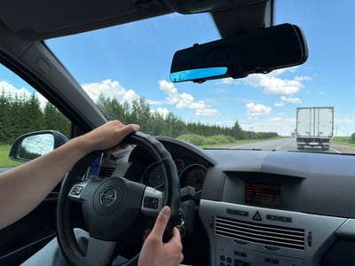 A first-person view from the driver's seat of a car shows a person's hands on the steering wheel. The car is driving on a road with trees on either side. A white truck is visible in the distance on the road ahead. The dashboard of the car displays information about the vehicle, including its speed, coolant temperature, and sensor readings. The sky is blue with some clouds. The scene is consistent with being on a highway or main road. The exact location is not specified in the image.