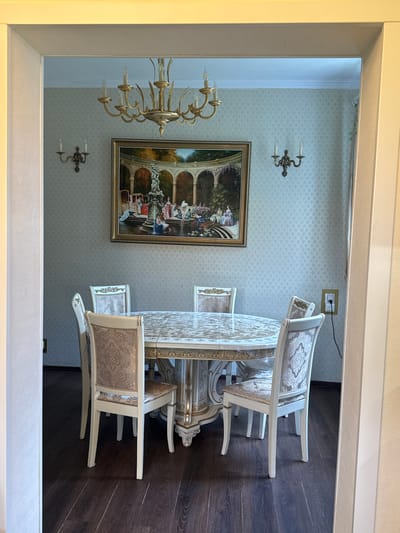 The image shows a dining room in a house in Vartemyaki, SNT, Russia. A round dining table with ornate detailing is set with a patterned tablecloth. Six chairs with upholstered seats and backs, featuring a light-colored fabric with a damask pattern, surround the table. The walls are covered in a light-colored wallpaper with a subtle, repeating pattern. A large, framed painting depicting a scene with figures in a garden setting hangs on the wall above the table. A chandelier with multiple arms and candle-shaped lights is suspended from the ceiling. Two wall sconces with candle-like lights are mounted on the walls. A power outlet with a black cord is visible on the right wall. The floor is dark wood laminate. The room appears to be well-lit, with light streaming in from an unseen source.