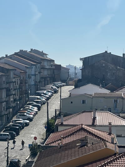 An aerial view captures a street lined with parked cars between tall buildings. The structures exhibit diverse architectural styles, with tiled roofs dominating the right side and modern apartments rising on the left. A few pedestrians are visible on the street. The sky is clear and bright.