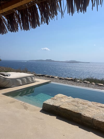 The image captures an outdoor private area in Agios Ioannis Diakoftis, Greece, featuring an infinity-edge swimming pool with an expansive view of the Aegean Sea. The upper portion of the frame is occupied by the underside of a straw or thatch roof overhang. A light-colored concrete or paved surface extends from the foreground, leading to the rectangular pool. To the left of the pool, a large, light-grey upholstered outdoor daybed rests on the patio. The pool water is clear blue, with visible submerged steps at the near end. A low wall of rough-hewn light brown stones forms the right border of the pool area. Beyond the paved area, a rocky and sparsely vegetated slope descends to the coastline. The Aegean Sea, characterized by deep blue, wind-rippled water, stretches to the horizon, dotted with several distant landmasses. The sky is clear and light blue, containing one small, isolated white cloud. A white vessel, possibly a speedboat, is visible on the water in the mid-left background, creating a distinct wake. No individuals are present in the scene.