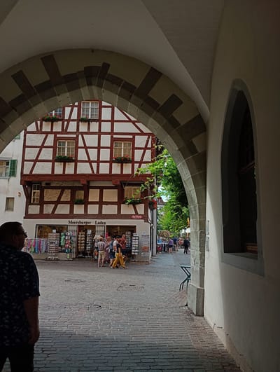 The image shows an archway in the city of Meersburg, Germany, framing a view of a cobblestone street lined with historic buildings. In the foreground, a man wearing a dark blue patterned shirt stands with his back to the camera. Through the arch, a half-timbered building with red trim and white infill is visible. Below the building, "Meersburger Laden" is written above a shop entrance. Several people are walking and standing on the street, some looking at merchandise displayed outside the shop. A green metal chair is visible to the right of the street. Lush green trees overhang the street in the background. The cobblestone pavement is worn and uneven.