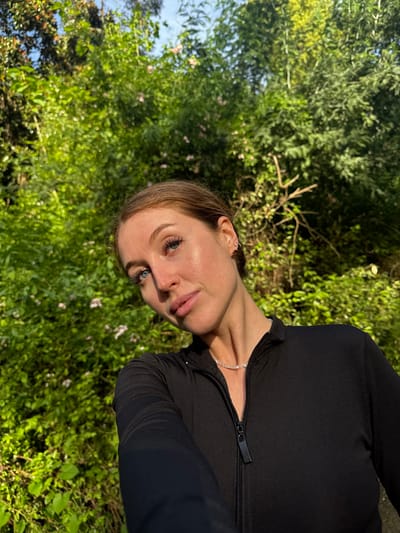 A woman is in a natural setting, likely a park or garden, possibly located in Tsanbaikwara, Abkhazia. She is wearing a dark-colored, possibly black, zip-up top. She appears to be fair-skinned with light brown hair and blue eyes. She wears a necklace. The background consists of dense greenery, with trees and bushes. The image appears to be taken from a slightly low angle.
