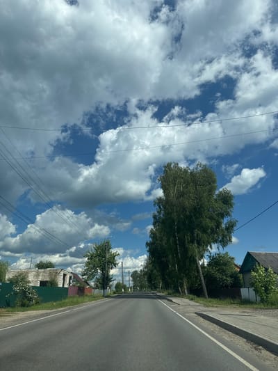 The image depicts a rural road in the village of Torbeyevo, Russia, on a cloudy day. The road is paved and has a white line marking the center. On the left side of the road, there is a green fence, a partially constructed building with white brick walls, and some trees with white flowers. Further down the road on the left, there are more trees and houses. On the right side of the road, there are several trees, including a tall birch tree, and a house with a dark roof. Power lines are visible overhead, crisscrossing the blue sky filled with dramatic white and gray clouds. The overall scene suggests a quiet, residential area in the countryside. There are no people visible in the image.