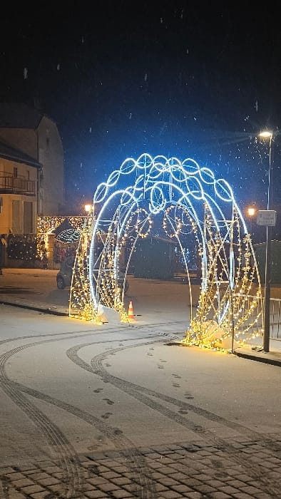 A wintery scene with a large, illuminated archway decorated with lights. The archway is constructed of metal and adorned with white and warm-toned lights. Tire tracks and footprints are visible in the snow-covered ground around the archway. A streetlamp and buildings are visible in the background. Snowflakes are apparent in the dark sky.