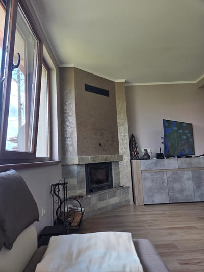 The image depicts a living room interior. On the left, a large window with wooden frames is partially open, revealing a view of the exterior. The interior features a fireplace with a stone surround, a modern entertainment unit with a large television screen displaying a map, and a sculptural decorative item. A basket filled with firewood is placed near the fireplace, along with a set of fireplace tools. A portion of a sofa with cushions is visible in the foreground. The walls are painted a neutral beige color, and the floor is covered with wooden laminate flooring. The lighting appears to be natural, coming from the window, and supplemented by artificial lighting.