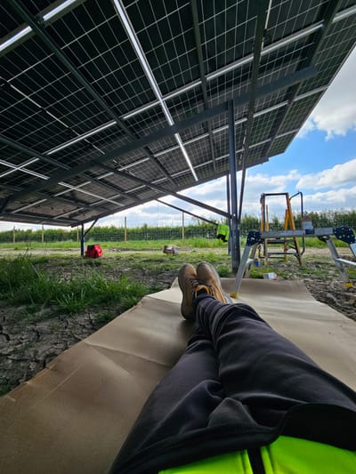 This image captures an outdoor scene at a solar panel installation or maintenance site, likely in Stroud, United Kingdom. The perspective is from a low angle, showing the lower body of a person resting while facing a large array of solar panels.
In the foreground, a person's legs are visible, clad in dark grey trousers and sturdy brown work boots, with their feet crossed. A bright neon yellow high-visibility vest is partially seen on their torso. They are resting on a large, unfolded piece of brown cardboard laid directly on the ground.
To the left, a significant structure of black solar panels extends upwards and to the left, showcasing their metallic support framework and grid patterns on the reverse side. The sky, a clear blue with scattered white clouds, is visible above the panels.
In the middle ground, beyond the resting person and the solar panels, various work-related items are present. A light-colored metal ladder or small scaffolding unit stands upright. A wire mesh fence runs horizontally across the landscape, separating the immediate foreground from a greener area. A red tool bag or container rests on the ground near the fence, and a small cardboard box is also visible. The ground itself is a mix of dry, cracked earth and patches of green grass.
The scene depicts a break from work during daylight hours. The weather appears fair and sunny with some cloud cover. No specific text is clearly legible on any objects in the image.