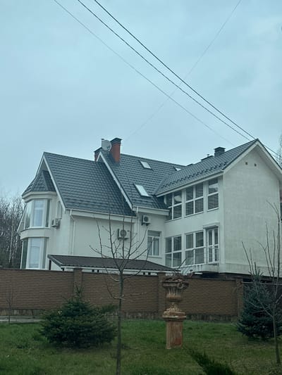The image shows a two-story residential building with a dark gray tiled roof and white walls, located in the city of Yurivka, Ukraine. The building has several windows, including a large bay window on the left side, and a patio covered by a smaller gray roof in front. There are two air conditioners on the exterior walls. The building is surrounded by a brown brick fence. There are three small trees in front of the fence, and a concrete sculpture in the form of an urn on a pedestal in the lawn. Several power lines run across the sky. The sky is overcast.  The location is 50.332 degrees latitude, 30.372 degrees longitude.