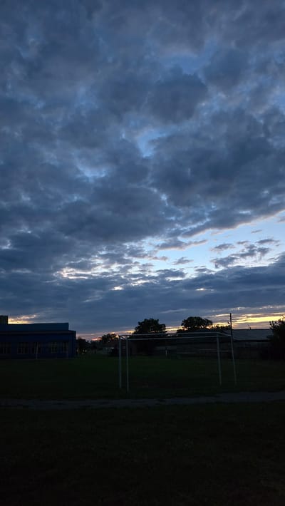 This is a vertical, low-angle, outdoor, daytime photograph taken at dusk in Alkhazurovo, Russia. The image captures a dramatic sky filled with dark, heavy clouds, interspersed with patches of lighter blue and hints of a sunset with orange and yellow hues peeking through on the horizon. The clouds are textured and appear to be moving, suggesting a dynamic weather pattern.
In the foreground, on the right side of the image, a dark, grassy field dominates the lower half. A gravel path runs along the edge of the field. Further back, a blue building with multiple windows is visible, partially obscured by trees. Several metal structures, possibly goalposts or playground equipment, are scattered across the field and near the building. The overall scene is dim, indicating the transition from day to night. The lighting suggests that the sun has set or is very low on the horizon. The mood is somewhat somber and atmospheric due to the heavy clouds and dim light. There is no visible text in the image.