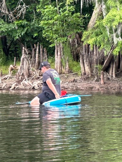An outdoor, slightly rotated image captures a man standing partially submerged in a body of water, interacting with a blue paddleboard or similar watercraft. The scene is set in a natural, lush environment, likely in Tampa, United States.
The man, facing left in the frame, wears a dark t-shirt with a green graphic design on the back, dark shorts, a dark baseball cap, and sunglasses. He is positioned with his legs in the water, appearing to be either preparing to launch or having just returned from an activity. He holds the blue and white watercraft with his hands. A red object, possibly a cooler or dry bag, is visible on the board.
The water is dark and reflective, showing streaks of light from the bright overhead sky. The background features a dense shoreline characterized by numerous exposed, gnarled tree roots and trunks extending into the water. Behind these roots, abundant green foliage and trees create a thick natural barrier, suggesting a riverine or swampy landscape. The overall impression is a clear, sunny day, ideal for outdoor recreational activities on the water. No distinct text is clearly legible on any visible objects.