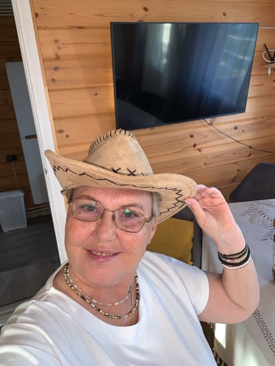 The image shows a woman wearing a light brown cowboy hat and glasses, with a white t-shirt and multiple beaded necklaces. She is also wearing bracelets on her left wrist. She is indoors, in what appears to be a room with wooden paneled walls. A large black television is mounted on the wall behind her. In the background, to the left, is a doorway leading to another room, where a white trash can is visible. To the right, a table with a patterned tablecloth is partially visible, along with a dark chair. The lighting suggests it is daytime. The location is identified as Суроватиха, Russia.