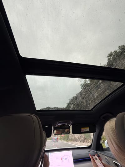 The image depicts the interior of a vehicle, possibly a car or SUV, on a cloudy, rainy day. Water droplets are visible on the windows, creating a blurred and streaky view of the outside environment. The interior shows the back of a passenger seat in the foreground, with dark trim and upholstery.
Looking towards the front of the vehicle, we see parts of the dashboard and ceiling. A sun visor with warning labels, including an airbag symbol, is visible on the driver's side. A rearview mirror is mounted below it, reflecting a glimpse of the road ahead and a distant car. To the right, a large touchscreen display is present, showing what appears to be a charging indicator with "25%" displayed. A person's hand with red nail polish is holding a smartphone near the screen. The hair of another person, with blonde highlights, is also visible in the upper right portion of the frame.
Outside the windows, a landscape of rocky hills and vegetation can be seen through the rain. The weather is clearly overcast, suggesting it might be daytime, but the lighting is subdued. The scene conveys a sense of travel and being inside a vehicle during inclement weather. The location is identified as Donji Ulići, Montenegro.