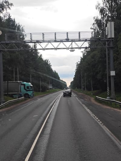 The image depicts a highway scene in Zhel'tsy, Russia, on an overcast day. A paved road with a white lane marker stretches into the distance, flanked by dense trees on both sides. Overhead, a metal gantry supports various equipment, including cameras and lights, likely for traffic monitoring or toll collection.
In the foreground on the left, a light blue truck with a silver trailer is parked on the shoulder, partially obscured by a metal guardrail. A person in a blue shirt is visible near the truck, appearing to be either working on or inspecting it.
Further down the road, a dark-colored passenger car is traveling away from the camera. The road surface appears wet, indicating recent rain. The environment suggests a rural or semi-rural highway setting.