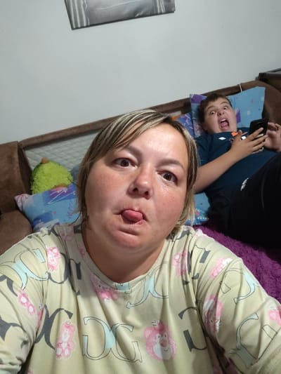 The image shows a selfie taken indoors, likely in a residential setting. The foreground features a woman sticking her tongue out. In the background, a boy is lying down on a sofa, looking at his phone with his mouth open in a surprised expression. He is wearing a dark blue t-shirt. The sofa appears to be brown, with colorful pillows. A fuzzy green stuffed toy is also visible on the sofa. The overall environment suggests a casual, home-like atmosphere. The image does not contain any information that would allow for the determination of its location in the city of Nazarovo, Russia, or any other specific geographical location.