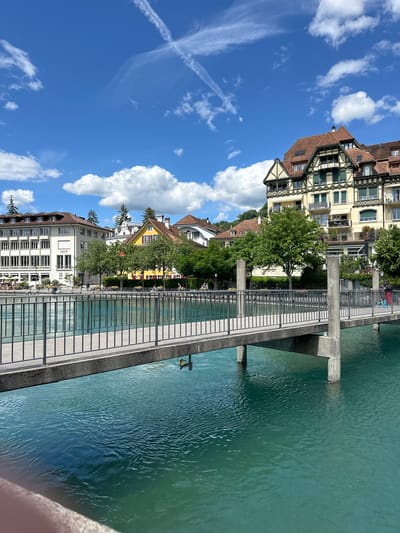 The image depicts a view of the Aare River in Thun, Switzerland, on a sunny day with scattered white clouds in a blue sky. A pedestrian bridge with a metal railing spans the turquoise water. On the left bank, several buildings are visible, including a modern white building with "AEK" signage. On the right bank, a large, multi-story building with a distinctive architectural style, featuring a red tiled roof and yellow facade, dominates the scene. Lush green trees line the riverbanks, and a few people can be seen walking on the promenade. The water is clear and reflective, with gentle ripples on its surface.