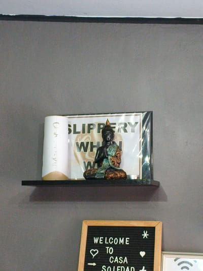 An indoor shelf displays a white cylindrical object with a gold base and a framed sign with a Buddha statue in front of it. The framed sign reads "SLIPPERY WHEN WET". Below the shelf, a black letter board with white text says "WELCOME TO CASA SOLEDAD". The scene is set against a gray wall, suggesting an interior space.
