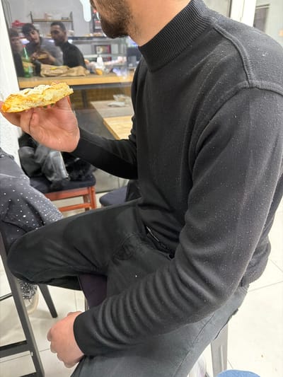 A person wearing a black sweater and pants is holding a piece of bread with a white topping. The scene is indoors, reflected in a window, and appears to be in the city of Strumica, North Macedonia, judging from the reflections in the window. The person is looking down at the bread, suggesting they might be examining or preparing to eat it. Other objects in the scene include a chair, a bag, and other items visible in the window reflection.