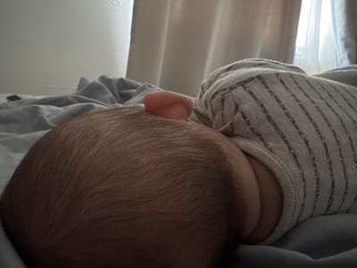 The image shows a close-up view of a baby's head and upper body, lying on a bed covered with grey and white striped fabric. The baby's head is to the left of the frame, with fine, light brown hair. A small, pink ear is visible on the side of the head, protruding slightly. The baby is wearing a grey and white striped garment. The background is out of focus, with hints of a white wall and curtains, suggesting an indoor setting. There is no identifiable information to place this scene in Oldham, United Kingdom.