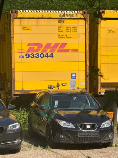 The image captures an outdoor scene in Schkopau, Germany, on a bright, sunny day. The central focus is a large, yellow shipping container with the DHL logo prominently displayed. Text on the container indicates it is GESU 704603 4, DMG1, and provides weight specifications for MGW, Tare, and Payload, along with the owner, GE SeaCo SRL, Bridgetown, Barbados. A warning sticker states "Caution 2.55m Wide".

In front of the container, two black cars are parked. The car on the right, a hatchback, is partially obscured by foliage. The car on the left, a sedan, is closer to the foreground. The ground is a gravelly surface with sparse vegetation. The background features green foliage and a glimpse of a structure, possibly another shipping container, to the right of the main container. The scene suggests a logistics or industrial area, possibly a storage yard or a transportation hub. There are no people visible in the image.