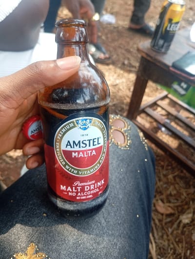 A close-up outdoor photograph depicts a dark-skinned hand holding an opened 330ml amber glass bottle of Amstel Malta, a non-alcoholic premium malt drink. The bottle contains a dark liquid with visible foam at the top, and its red crown cap, bearing the "AMSTEL MALTA" logo, is held between two fingers of the same hand. The bottle rests on the upper thigh of an individual wearing dark denim jeans embellished with gold-colored studs. To the right, in the midground, a small, dark wooden table holds a black and yellow can of Legend Extra Stout and a dark-cased smartphone. The ground is reddish-brown earth, covered with scattered dry leaves. Several blurred figures are visible in the background, partially showing legs and feet in various casual footwear, including sandals and dark shoes, consistent with an outdoor social gathering. The scene is illuminated by natural daylight and is located in Egede, Nigeria.