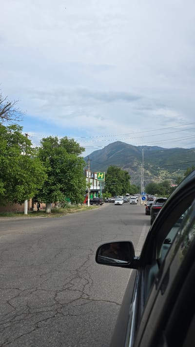 A road scene is depicted, likely within the city limits of Charvak, Uzbekistan, under a partly cloudy sky. The foreground is dominated by the side of a dark-colored vehicle, including its side mirror which reflects a person's face. The road surface is asphalt, showing signs of wear and tear with visible cracks. To the left of the road, a sidewalk and a building are present, partially obscured by lush green trees. Further down the road, a line of vehicles is moving, and on the right side, there is a large green sign with a white "H" and "HAVAS" written in white. In the background, a mountain range rises, covered in green foliage, suggesting a mountainous or hilly terrain. Power lines are visible crisscrossing the sky. A small, dark object, possibly a bird or drone, is suspended high in the sky.