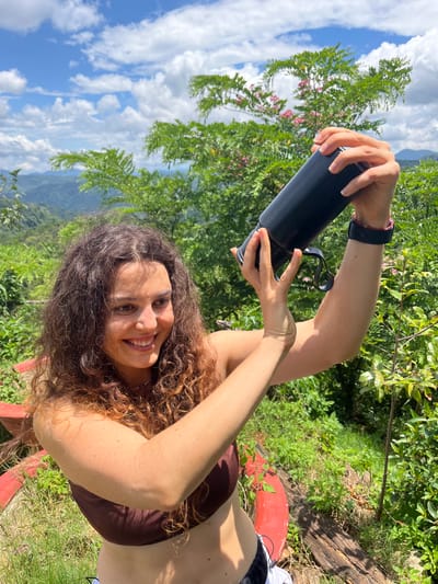 A young woman with curly brown hair and a brown bikini top is taking a selfie with a black water bottle held in her hands. She is outdoors in a lush, green environment with mountains visible in the background under a blue sky with white clouds. The woman is smiling at the camera. The location is identified as Sơn Điền, Vietnam.