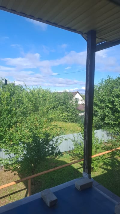This image captures a view from a covered porch or balcony, looking out onto a residential area on a clear, sunny day. The foreground shows a dark blue surface with two concrete blocks resting on it, possibly used as weights. A rust-colored railing runs horizontally across the mid-ground, casting a shadow. Beyond the railing, a corrugated metal fence encloses a yard filled with lush green trees and vegetation. In the background, a white house with a brown roof is visible through the foliage, suggesting a suburban or rural setting within Engels, Russia. The sky above is a vibrant blue, dotted with scattered white clouds. The overall scene depicts a peaceful, everyday outdoor environment.