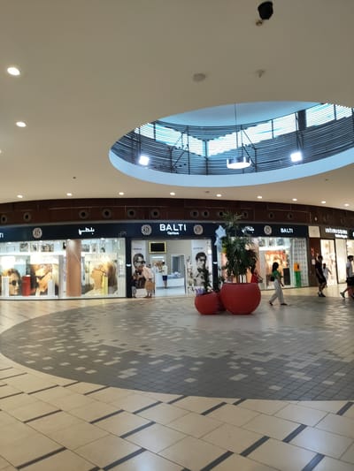 This image depicts the interior of a modern shopping mall, likely in Cebalat, Tunisia. The scene focuses on a wide, tiled walkway leading towards a row of shops. The shops feature large display windows showcasing various products, including eyewear, as evidenced by prominent "BALTI OPTIQUE" signage and images of sunglasses. One shop is labeled "Intimissimi," suggesting lingerie or apparel. The mall has a high, domed ceiling with a circular opening that allows natural light to filter in, supplemented by artificial lighting. Several people are visible walking through the mall, some browsing in front of the shops, while others are in motion along the walkway. The floor has a distinctive tiled pattern, with a large circular area of darker, pixelated tiles. Large, red decorative planters with greenery are placed in front of some of the shops, adding a touch of color to the environment. The overall impression is that of a clean, well-lit, and contemporary retail space.