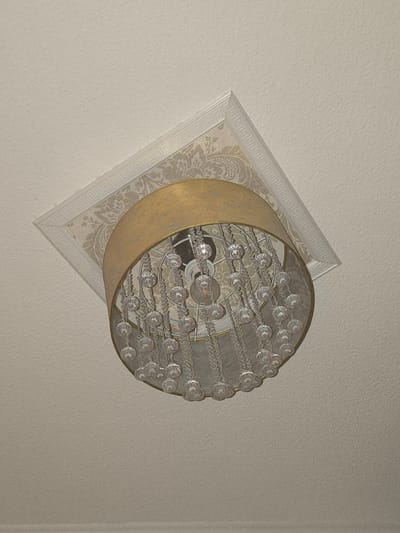 The image shows a close-up, slightly angled view of a chandelier hanging from a textured, off-white ceiling. The chandelier features a gold-colored drum shade and is adorned with numerous clear, faceted crystal beads that hang in multiple strands. The ceiling has a decorative cornice, with a Greek key pattern along the outer edge and a floral damask pattern closer to the chandelier. The lighting appears to be soft, with the light bulb inside the shade partially visible. The scene is indoors, likely in a residential setting, and the location in Bautzen, Germany is not discernible from the image itself. There are no people or specific actions depicted.