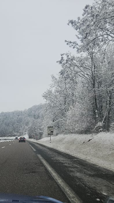 The image depicts a winter scene on an asphalt highway near Thionville, France, under an overcast grey sky. The road features at least two visible lanes with faded white dashed markings. Snow and slush partially cover the right shoulder and median. A dark-colored sedan with illuminated red brake lights is observed in the distance, traveling in the left lane. The perspective is from inside a vehicle, with a portion of its blue dashboard visible in the lower foreground.
To the right of the road, a snow-covered embankment slopes upwards, bordered by a dense forest of deciduous trees heavily laden with snow. A black and white rectangular road sign stands on the right, displaying symbols for slippery conditions (ice/snow), various vehicle types (car, truck, trailer), and possibly lane or distance regulations. The word "Thionville" is legible on the sign. Further into the background on the left, a snow-covered, forested hill is visible. The entire environment is characterized by significant snowfall and winter conditions.