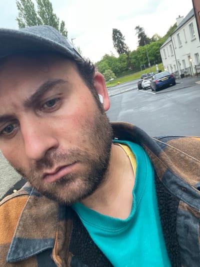 A close-up, slightly angled portrait of a bearded man wearing a gray baseball cap, a teal shirt, and a brown and navy plaid jacket. The background shows a street in Armagh, United Kingdom with a white building, cars, and greenery. The man is the primary subject, occupying the foreground. The focus is primarily on his face.