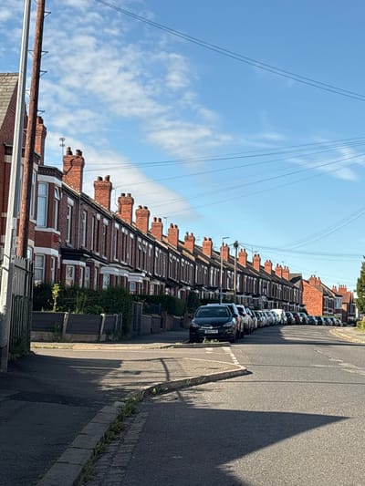 The image depicts a residential street in Crewe, United Kingdom, lined with terraced houses. The houses are constructed of red brick with white-trimmed windows and prominent chimneys. A row of parked cars, mostly dark in color, occupies the side of the road. The sky is blue with scattered white clouds. A tall wooden pole with metal attachments, likely a utility pole, stands on the left side of the image, in front of the houses. The road surface is asphalt with a distinct curb and a paved sidewalk or verge. The scene is illuminated by sunlight, casting shadows on the road. The overall impression is a typical suburban street scene on a clear day.