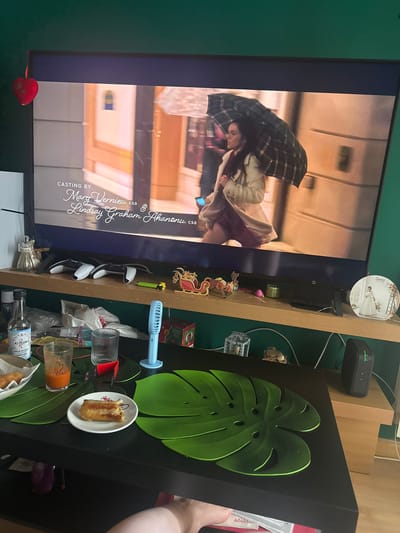 This media file is a photograph taken in a room, likely a living space or entertainment area, with a large television screen dominating the left side of the frame. The television displays a scene from a movie or show featuring a woman holding a large, dark plaid umbrella over herself. She appears to be indoors, possibly in a hallway or lobby, with blurred background elements suggesting a modern architectural setting with light-colored walls and possibly glass.

To the right of the television, a wooden shelf unit is visible, cluttered with various objects. Among these are a small blue fan, several decorative items including what looks like a small Christmas ornament with a reindeer, and a round decorative piece with a picture of a woman in a white dress. There are also some electronic devices and wires. Below the shelf, a black surface is covered with more items, including two pieces of food on a white plate, glasses, bottles, and a large green monstera leaf-shaped placemat or decoration. The overall impression is of a casual, lived-in environment.

The lighting suggests it is daytime, but the indoor setting makes it difficult to ascertain the time of day or specific weather conditions. The presence of a movie on the screen and the scattered items on the shelf imply a relaxed, possibly home-viewing scenario. The text on the TV screen provides casting information: "CASTING BY Marg Vernieu, csa & Lindsay Graham Ahanonu, csa". There is also text on a bottle on the right, which appears to be "SALSA DE SOJA" (Soy Sauce in Spanish) and possibly "KIMONJ" in smaller print. The location context provided indicates Alcalá De Henares, Spain, which aligns with the Spanish text on the bottle.