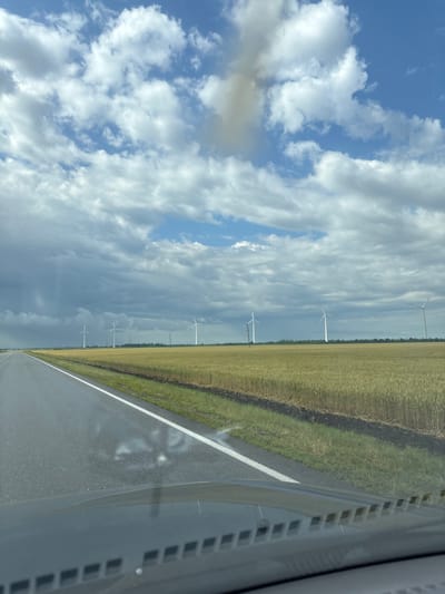 The image depicts a wide, asphalt road stretching into the distance. A white line marks the right edge of the road. To the right of the road, there is a field of dry, golden grass, likely a harvested crop. Beyond the field, a line of white wind turbines stands against a dramatic sky. The sky is a mixture of bright blue with fluffy white clouds and darker, overcast areas with patches of blue peeking through. The clouds suggest a dynamic weather system, possibly with impending rain or recent showers. The scene appears to be set in a rural, open landscape. The perspective is from inside a vehicle, with the dashboard and windshield visible in the lower right corner. The light suggests it is daytime, likely late morning or afternoon. There is no visible text in the image. The overall mood is one of openness and the vastness of nature, with the human-made structures of the road and wind turbines interspersed within the landscape.