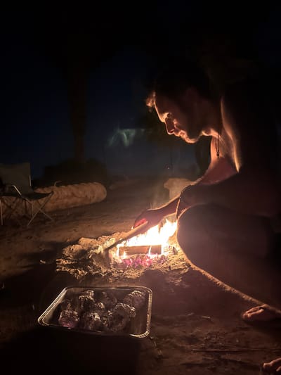 The image depicts a nighttime outdoor scene, presumably within the Arava Tichona Regional Council, Israel, illuminated primarily by a campfire. A male individual, shirtless with a dark pendant necklace and short dark hair, is shown in profile, squatting or kneeling on the right side of the frame, intently focused on the fire. He extends his right hand, holding a light-colored stick or piece of wood into the active flames. The campfire, contained within a small ring of rocks and disturbed earth, displays glowing embers and bright flames.
To the left of the fire, in the foreground, a rectangular aluminum foil tray contains multiple items individually wrapped in aluminum foil, presumably food, ready for cooking near the fire's heat. In the background, to the far left, a light-colored foldable camping chair is visible, and behind it, a long, thick log rests horizontally on the sandy ground. A dark, indistinct tree trunk or structure is silhouetted against the dark night sky in the distant background. The ground is consistently sandy or dirt-covered.